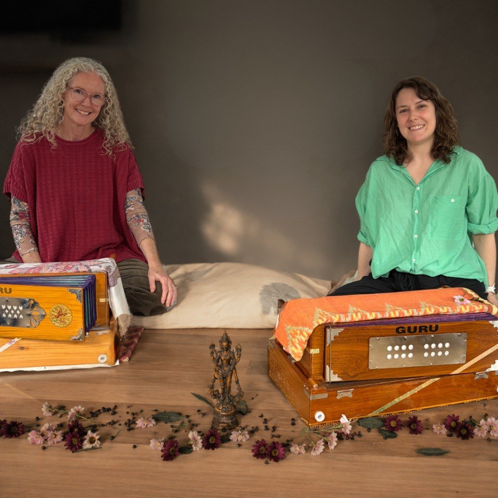 Sharon and Erin sitting comfortably with beautiful wooden harmoniums, scattered flowers around them, in a warm intimate studio setting for sound meditation practice